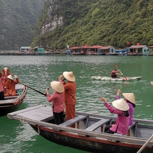 Local rowers in conical hats waving from small boats on green water near a floating fishing village